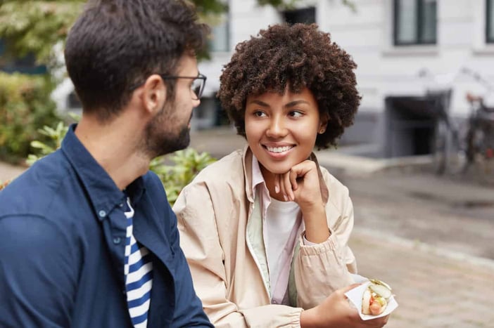 Photo of mixed race tourist enjoy lively communication, discuss traditions of their countries, eat fastfood on street, being in good mood. Beautiful Afro American girl talks with handsome guy outdoor