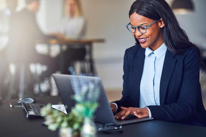 Smiling young African American businesswoman working on a laptop at her desk in a bright modern office with colleagues in the background Smiling young African American businesswoman working on a laptop at her desk in a bright modern office with colleagues in the background
