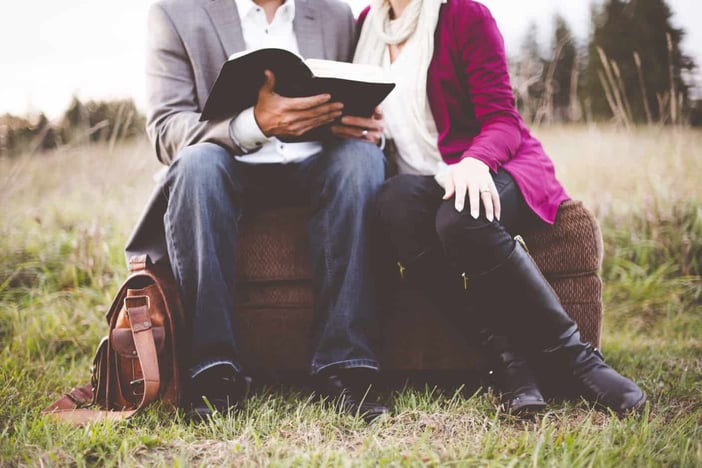 A closeup shot of a couple sitting on a suitcase while reading the bible with a blurred background