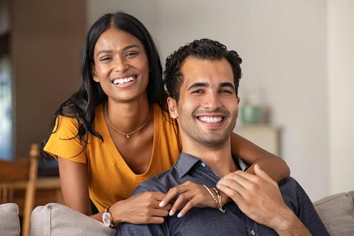 Smiling indian woman hugging her husband on the couch from behind at home. Loving middle eastern couple looking at camera with big grin. Portrait of laughing girl embracing handsome latin man on sofa.