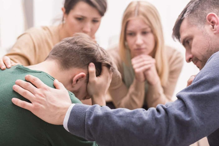 Close-up of a devastated young man holding his head in his hands and friends supporting him during group therapy Close-up of a devastated young man holding his head in his hands and friends supporting him during group therapy