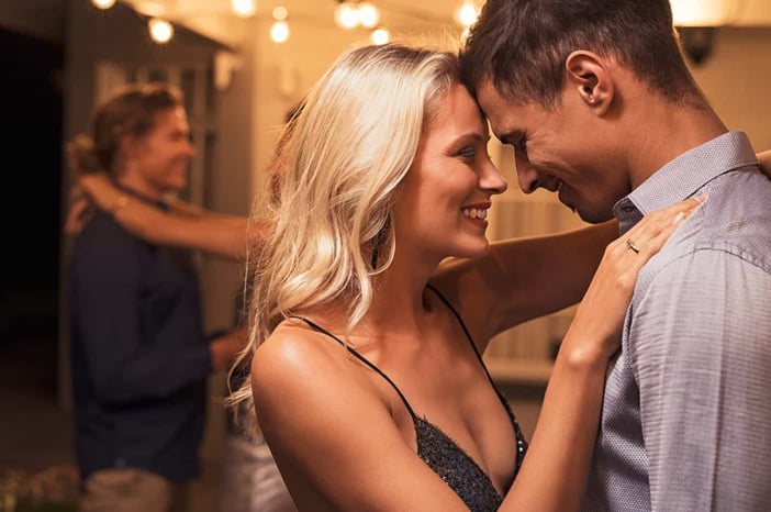 Beautiful loving couple dancing under the patio illuminated by wires of light bulbs. Young woman in love hugging her boyfriend. Romantic girl and elegant guy embracing outdoor at party night.