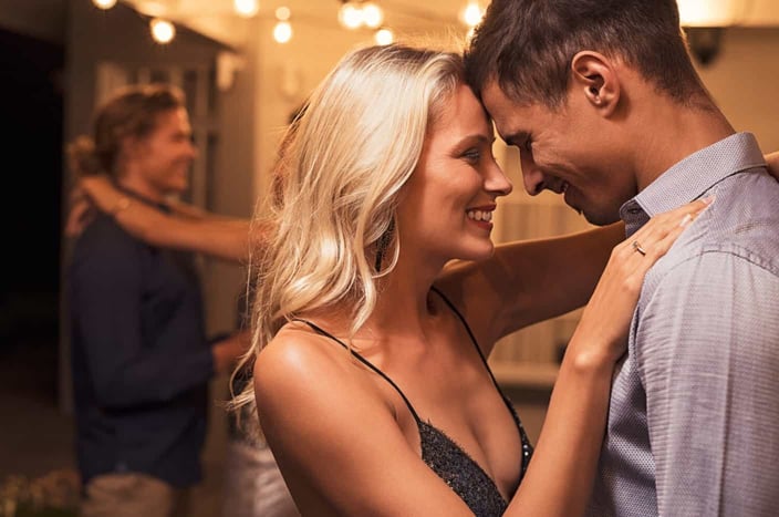 Beautiful loving couple dancing under the patio illuminated by wires of light bulbs. Young woman in love hugging her boyfriend. Romantic girl and elegant guy embracing outdoor at party night.