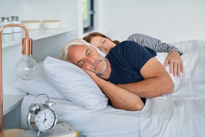 Senior man and mature woman sleeping together in their bed. Married middle aged couple resting with eyes closed in the morning. Wife lying on side embracing her husband while dreaming. 