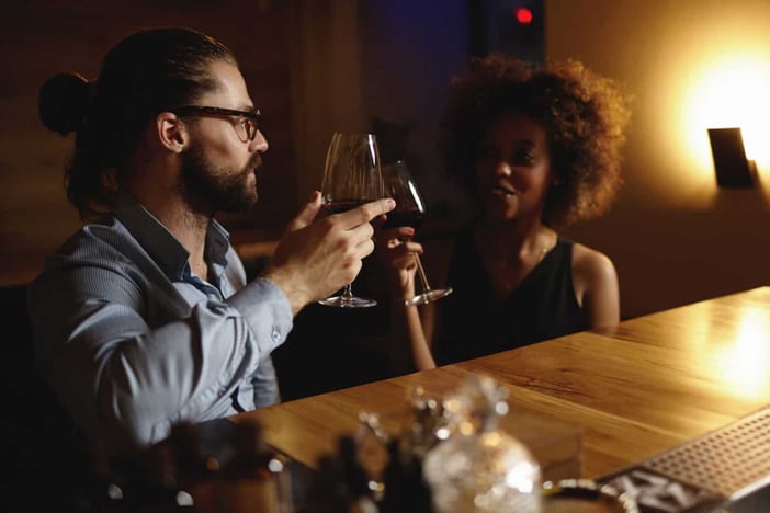 Fashionable interracial couple drinking wine during date, sitting at restaurant, having romantic evening and nice conversation, raising glasses to love at first sight. Hipster man proposing toast