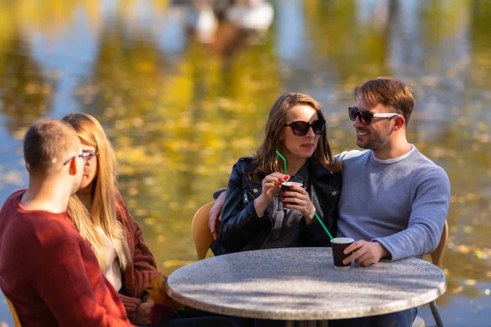 Two young couples enjoying coffee outdoors seated at a restaurant table overlooking tranquil water with autumn reflections