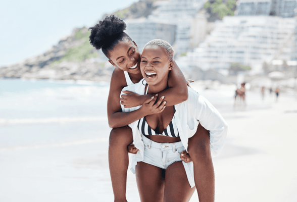 Two black lesbian women walking piggyback on a beach