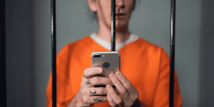 Prison inmate in orange jumpsuit with hand tattoos holding smartphone through cell bars Prison inmate in orange jumpsuit with hand tattoos holding smartphone through cell bars