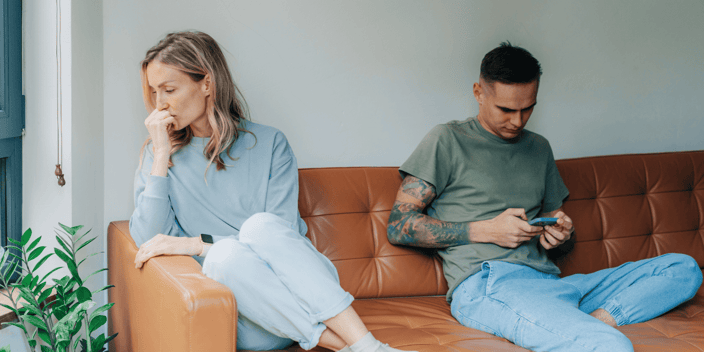 Man and woman couple with man on cellphone and woman upset sitting away from him on couch
