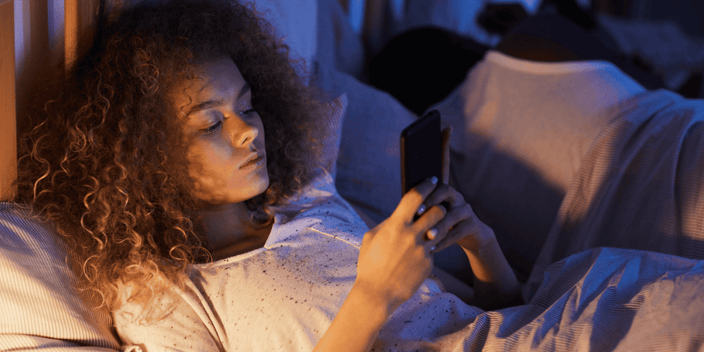 Young woman with curly hair scrolling on her phone in bed next to her sleeping boyfriend Young woman with curly hair scrolling on her phone in bed next to her sleeping boyfriend