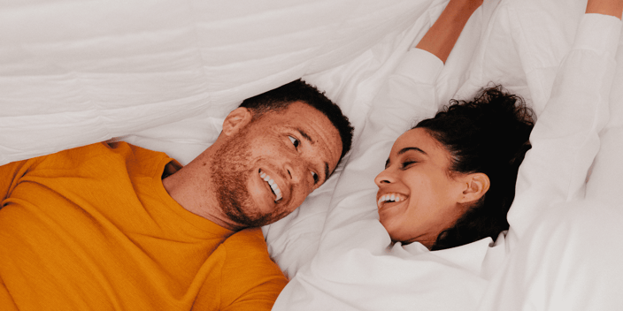 Young couple laying together under white bedsheet and laughing