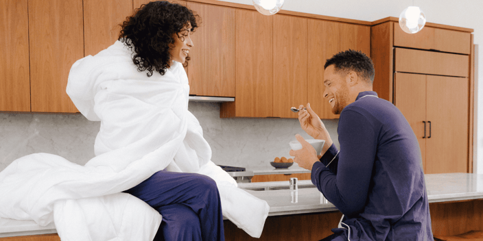 Young black couple sitting at kitchen counter laughing and having breakfast, woman wrapped in white comforter