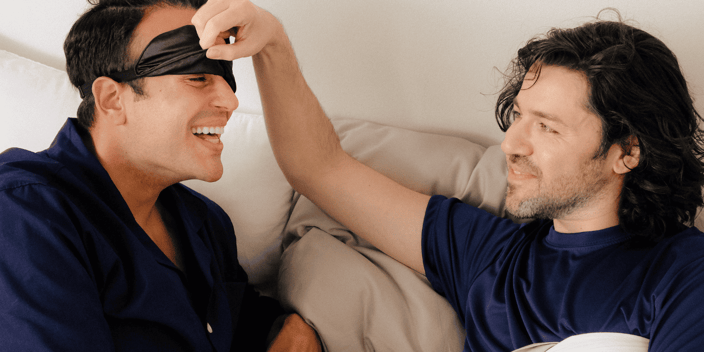 Two gay men in bed with one lifting the sleep mask off of the other