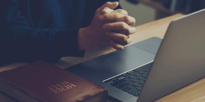 Man sitting with hands in prayer in front of his laptop and Bible