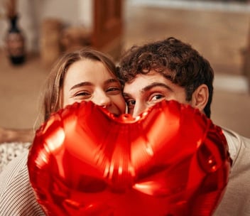 Photo of a male and female couple smiling behind a red heart balloon Photo of a male and female couple smiling behind a red heart balloon