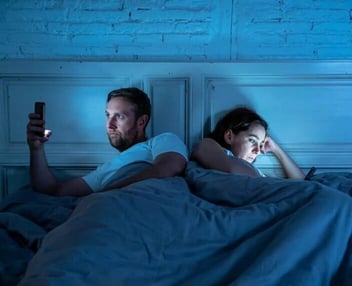 A photo of a couple facing in opposite directions in bed and staring at their cell phones