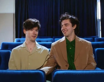 Screenshot from It's Funny We Met On Hinge commercial of a male couple sitting in a movie theater and smiling Screenshot from It's Funny We Met On Hinge commercial of a male couple sitting in a movie theater and smiling