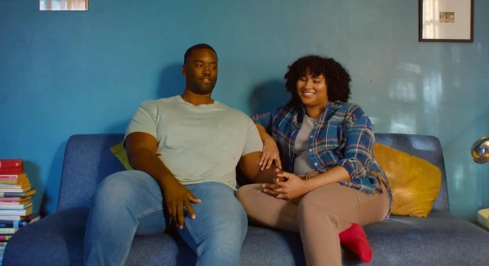 Screenshot from It's Funny We Met On Hinge commercial of a young Black couple sitting on a couch and holding hands and smiling Screenshot from It's Funny We Met On Hinge commercial of a young Black couple sitting on a couch and holding hands and smiling