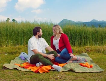 Screenshot from It's Funny We Met On Hinge commercial of a male and female couple sitting on a picnic blanket outdoors and smiling Screenshot from It's Funny We Met On Hinge commercial of a male and female couple sitting on a picnic blanket outdoors and smiling