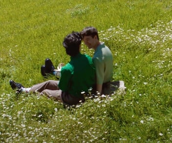 Screenshot from It's Funny We Met On Hinge commercial of two men sitting in a grassy field and smiling at each other Screenshot from It's Funny We Met On Hinge commercial of two men sitting in a grassy field and smiling at each other