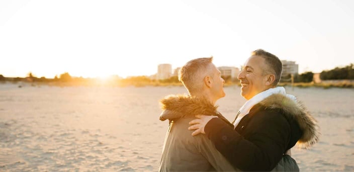 Two men in parkas holding one another by the ocean during sunset. Two men in parkas holding one another by the ocean during sunset.