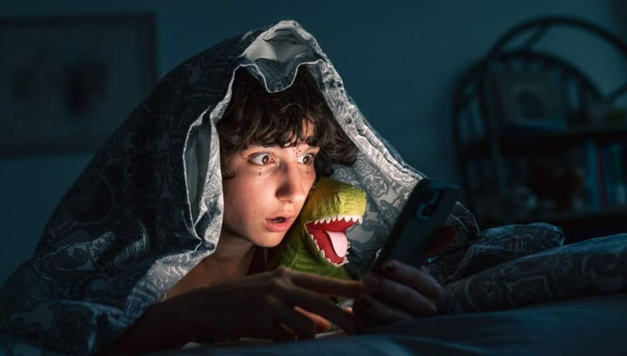A tween child, hiding under their blanket at night, looks shocked while staring at a cellphone screen A tween child, hiding under their blanket at night, looks shocked while staring at a cellphone screen