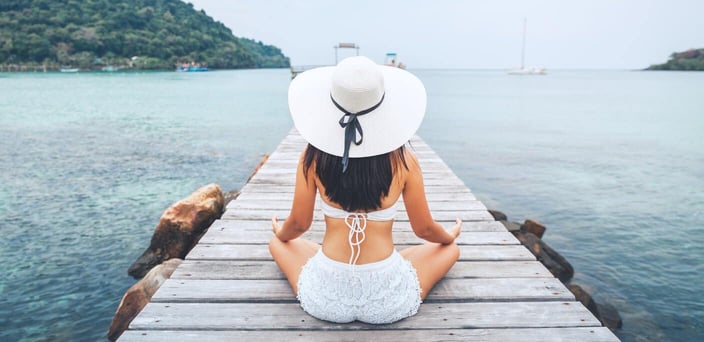 Woman in white hat and bathing suit sitting on pier in tropical location.