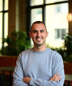 Michael Kaye smiles with his arms crossed while wearing a light blue crewneck shirt in a softly lit indoor setting. Michael Kaye smiles with his arms crossed while wearing a light blue crewneck shirt in a softly lit indoor setting.