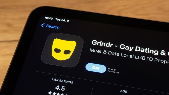 Grindr app listing on a mobile phone with a blurred rainbow flag in the background. Grindr app listing on a mobile phone with a blurred rainbow flag in the background.